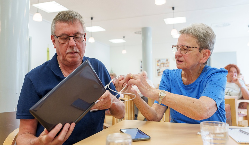 Eine ältere Dame hilft einem älteren Herren mit seinem Laptop.